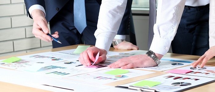 Auf einem Tisch liegen Projekte. 3 Personen befinden sich in einer Prozessberatung. Es werden Post-It und Istogrammen gezeigt.